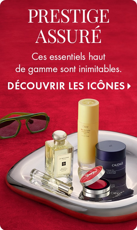 « PRESTIGE ASSURÉ » « Ces essentiels haut de gamme sont inimitables. » « MAGASINER LES ICÔNES » Essentiels beauté de prestige présentés sur un plateau argenté réfléchissant sur un fond rouge, y compris un parfum Jo Malone, des soins pour la peau, un boîtie