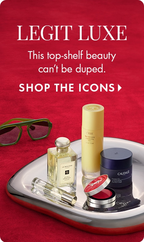 "LEGIT LUXE" "This top-shelf beauty can’t be duped." "SHOP THE ICONS" Luxury beauty products displayed on a reflective silver tray against a red background, including Jo Malone fragrance, skincare items, makeup compact, and green sunglasses.