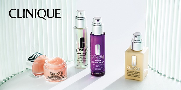 Image shows four Clinique skincare products on a bright white surface: an open peach cream jar, two pump serums in silver and purple bottles, and a beige moisturizer pump bottle, with the Clinique logo above, lit by soft vertical light reflections.