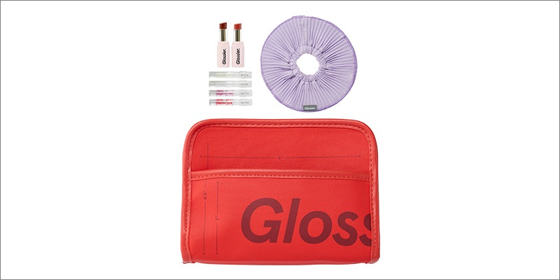 A red Glossier-branded zip pouch sits in the foreground. Above it are two Glossier lip products, three clear Glossier skincare mini bottles, and a light purple scrunchie-style hair accessory.