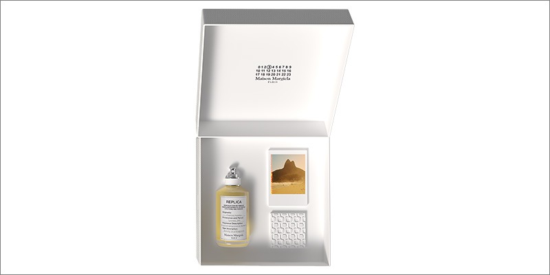 Maison Margiela Replica fragrance set displayed in an open white box, featuring a glass perfume bottle, a small photo card with a desert landscape, and a textured white accessory insert.