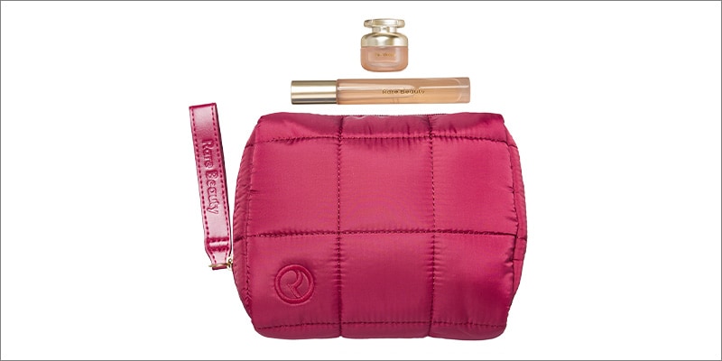 A magenta quilted Rare Beauty makeup pouch with a wrist strap and embossed logo. Above the pouch are two Rare Beauty makeup products: a small glass jar and a liquid concealer tube with gold-toned caps, arranged on a white background.