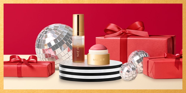 MERIT beauty products displayed on stacked platforms with red gift boxes and silver disco balls on a bold red background for a festive, polished look.