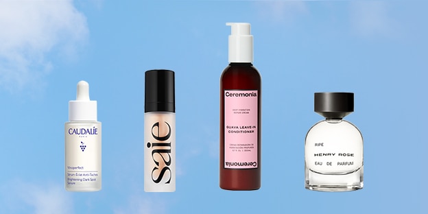 Four beauty products are arranged in a row against a light blue sky background: a small dropper bottle, a tall foundation bottle, a pink cylindrical bottle, and a round clear perfume bottle with a black cap.